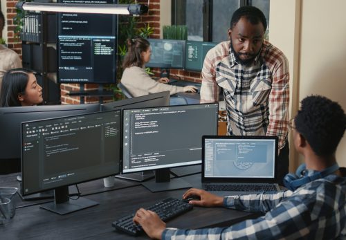 programer-sitting-desk-with-multiple-screens-running-code-talking-with-colleague-about-artificial-intelligence-algorithm-software-developers-doing-innovative-artificial-intelligence-project-scaled-1